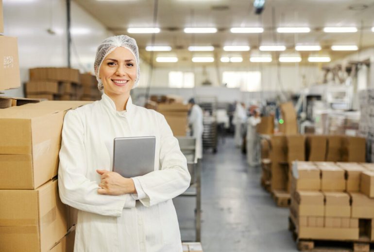 Portrait of food factory inspector leaning on boxes with merchandise and smiling at the camera.